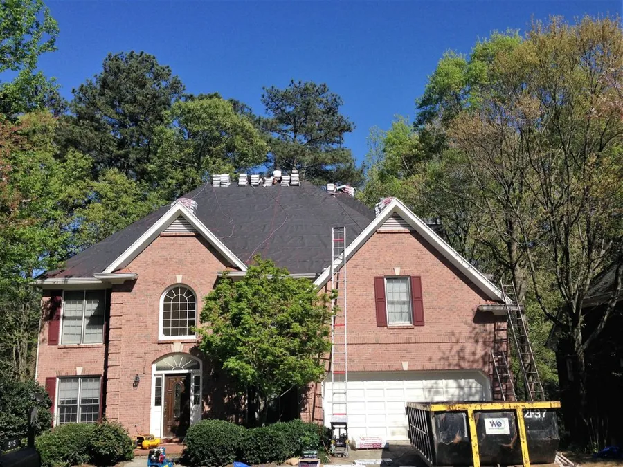 Finished dark shingle roof on a two-story brick home, with the tear-off dumpster still on site