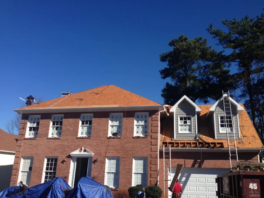 Brick colonial stripped down to new roof decking during a full tear-off, with the shrubs covered by tarps